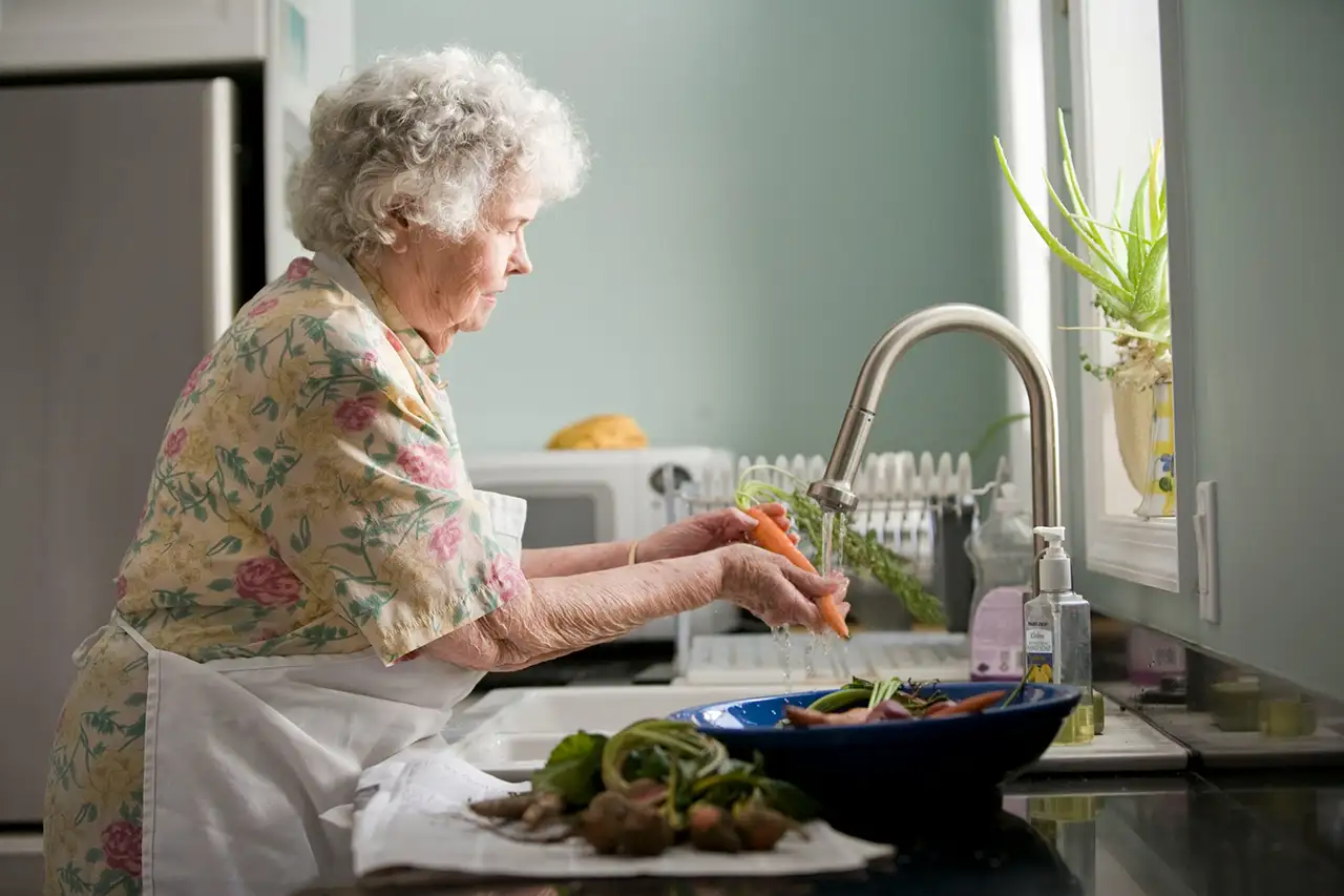Elderly woman washing vegetables in her kitchen at home - monitoring elderly parents living alone in Australia